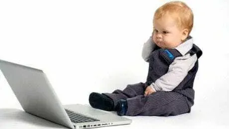 A toddler in a waistcoat sitting next to a laptop, looking puzzled